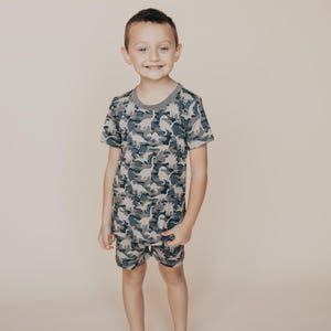 May include: A child wearing a matching two-piece pajama set. The set features a camouflage pattern with dinosaur silhouettes in shades of gray, blue, and white. The short-sleeved top has a gray neckline, and the shorts have an elastic waistband.