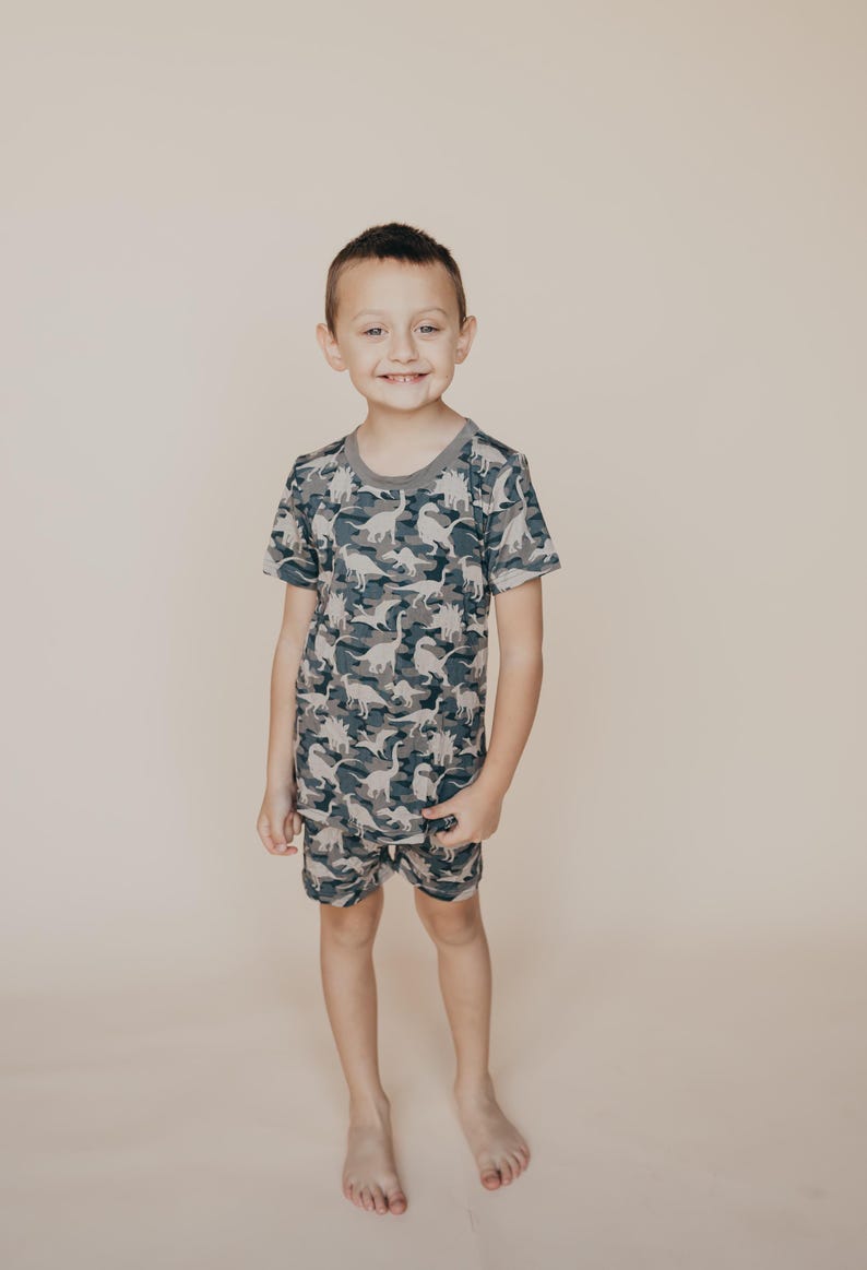 May include: A child wearing a matching two-piece pajama set. The set features a camouflage pattern with dinosaur silhouettes in shades of gray, blue, and white. The short-sleeved top has a gray neckline, and the shorts have an elastic waistband.