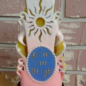 May include: A pink and yellow carved candle with a sun design and the words "At last I see The light" on a blue circle with white pearl accents.
