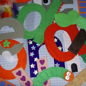 May include: A colorful felt craft project featuring large, whimsical letters, a green caterpillar, and various decorative elements. The letters are in shades of gray, green, and red, with googly eyes and star accents. The background is a blue and white checkered pattern.