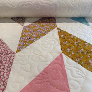 May include: Close-up of a quilted textile with a geometric pattern. The quilt features white, pink, and floral patterned fabric sections. The white sections have a floral design stitched into them. The quilt is rolled up at the top.