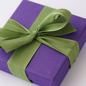 Puede incluir: Una caja de regalo morada con una cinta de sat&eacute;n verde atada en un lazo.