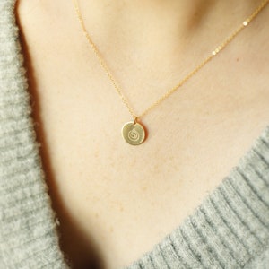 Puede incluir: Un collar de cadena de oro con un peque&ntilde;o colgante redondo con un dise&ntilde;o simple. El colgante cuelga del pecho de una persona.