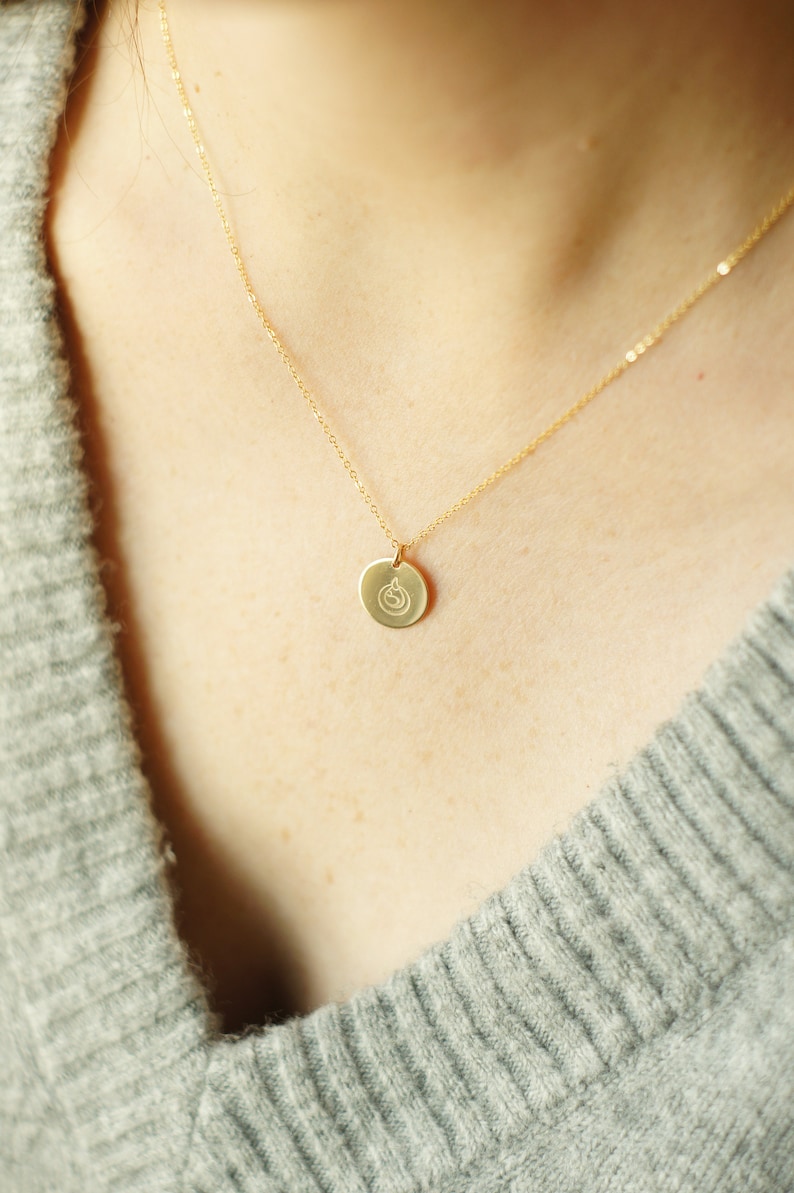 Puede incluir: Un collar de cadena de oro con un peque&ntilde;o colgante redondo con un dise&ntilde;o simple. El colgante cuelga del pecho de una persona.
