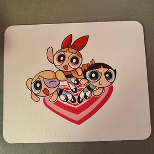 May include: A white square mousepad featuring the Powerpuff Girls cartoon characters. The image shows Blossom, Bubbles, and Buttercup in a dynamic pose over a pink heart design. The characters are in their signature colors.