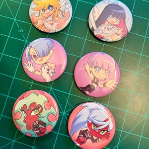 Puede incluir: Seis botones redondos con personajes de estilo anime. Los botones tienen fondos rosas, azules y blancos con ilustraciones coloridas de los personajes. Los personajes tienen alas de ángel, halos y otros accesorios.