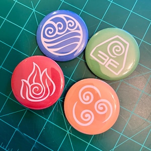 May include: Four round buttons with white symbols representing the elements of water, earth, fire, and air from Avatar: The Last Airbender. The buttons are blue, green, red, and orange, and are on a green cutting mat.