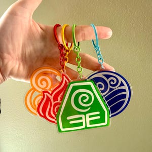 Peut inclure: Ensemble de porte-clés colorés représentant les éléments feu, terre, eau et air. Chaque porte-clés présente un design unique et est attaché à une chaîne avec un anneau de couleur différente.