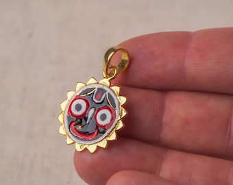 May include: Gold pendant featuring a black and red image of a deity with white eyes. The pendant is shaped like a sun with gold rays.