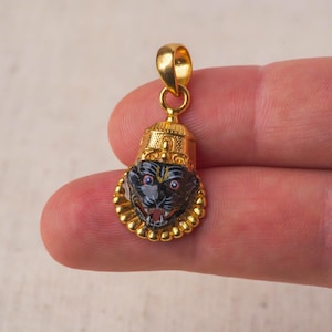 Op de afbeelding: Gouden hanger met een zwarte emaille afbeelding van een grommend dierenhoofd. De hanger is versierd met gouden kralen rond de rand.