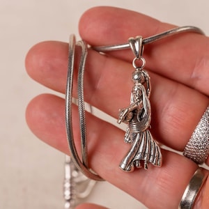 May include: A silver pendant necklace with a detailed figure in traditional dress. The pendant is suspended from a fine silver chain. Also visible is a silver ring with a textured surface.