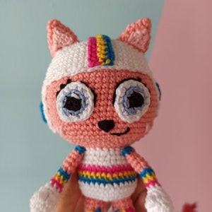 Puede incluir: Muñeca de gato de ganchillo hecha a mano con cuerpo rosa, gorro blanco y detalles azules y arcoíris. La muñeca tiene ojos azules grandes, nariz negra y boca sonriente. El gato lleva un casco blanco con una raya arcoíris.
