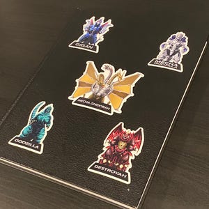 May include: Five colorful stickers featuring different Godzilla monsters. The stickers are on a black notebook. The stickers feature Godzilla, Gigan, Mecha Godzilla, Mecha Ghidorah, and Destroyah.