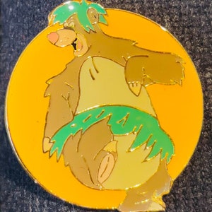 May include: A yellow and green enamel pin featuring a cartoon bear wearing a green grass skirt and a green leafy crown. The bear is smiling and has a pink nose.