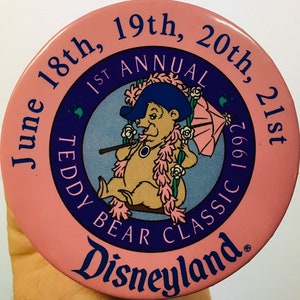 Könnte beinhalten: Ein runder rosa Knopf mit einem blauen und weißen Kreis in der Mitte. Der Kreis zeigt einen Comic-Teddybär, der einen blauen Hut trägt und einen rosa Regenschirm hält. Der Text um den Kreis lautet "June 18th, 19th, 20th, 21st 1st Annual Teddy Bear Classic 1992 Disneyland."