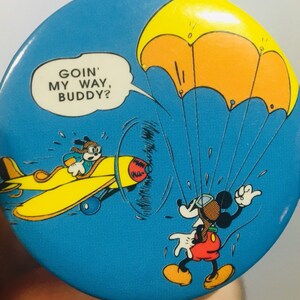 Op de afbeelding: Een ronde, blauwe knoop met stripfiguren. Mickey Mouse parachuteert, terwijl Goofy in een geel vliegtuig vliegt. Tekst in een tekstballon luidt: "Goin' my way, buddy?"