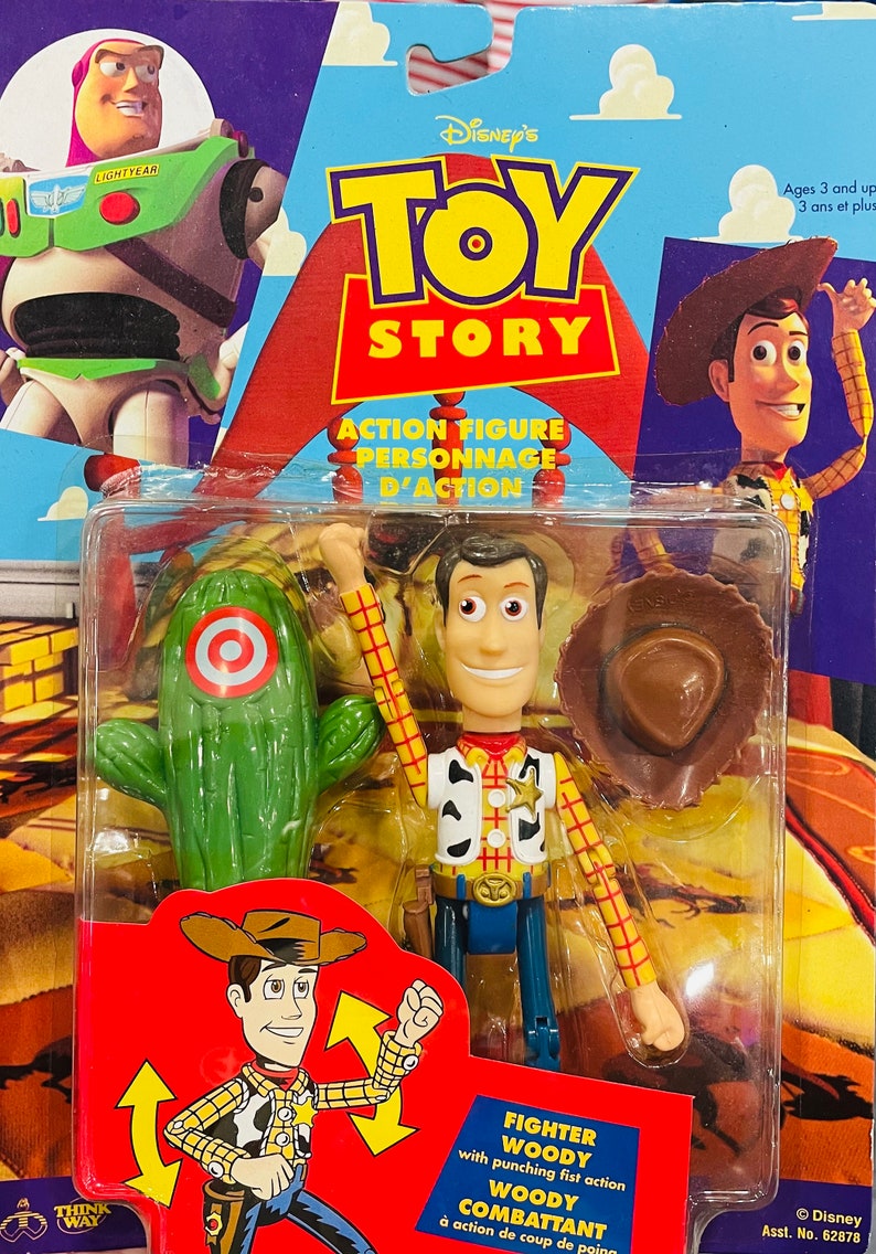 May include: Toy Story action figure of Woody, the cowboy doll, with a punching fist action. He is wearing his signature red and yellow plaid shirt, brown hat, and blue jeans. He is standing next to a green cactus with a red target on it. The packaging is red and blue with the Toy Story logo and the words "Action Figure" and "Personnage d'Action".