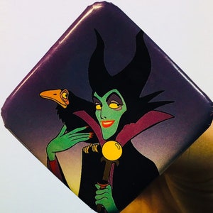 Op de afbeelding: Een vierkante knop met een cartoon afbeelding van Maleficent uit Disneys Doornroosje. Ze houdt een staf vast met een gele bol en een zwarte raaf zit op haar arm. De knop is paars, zwart en groen.