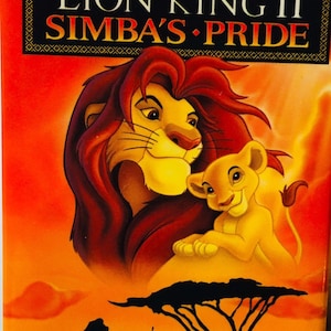 Puede incluir: Una carátula de cinta VHS para la película de Disney "El Rey León II: El reino de Simba". La carátula presenta un fondo rojo-naranja con una silueta de un árbol y un grupo de leones. Simba y su hijo, Kopa, están en primer plano, con Simba mirando al espectador y Kopa mirando a Simba. El texto "Disney's The Lion King II Simba's Pride" está en letras grandes y negritas en la parte superior de la carátula. El texto "Only On Video - October 27!" está en la parte inferior de la carátula.