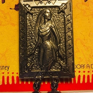 Puede incluir: Un pin de metal plateado con una imagen detallada de Pocahontas de la película de Disney. El pin está enmarcado con un borde decorativo y tiene tres hojas colgantes.