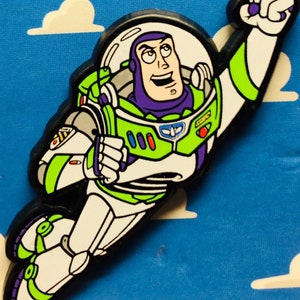 Puede incluir: Una figura de acción de Buzz Lightyear verde y blanca con un casco morado y un traje espacial negro. La figura está volando con un brazo levantado y una expresión decidida en su rostro.