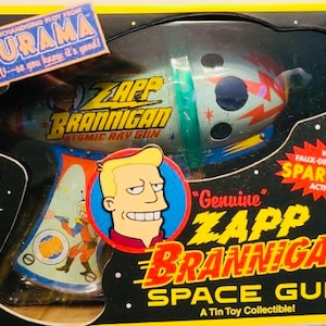 May include: A yellow and black box with a cartoon image of a space gun and a character from the animated TV show Futurama. The text on the box reads "Zapp Brannigan Space Gun" and "A Tin Toy Collectible!"