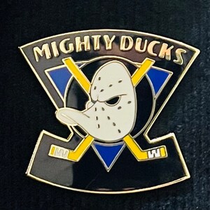 May include: A gold and black enamel pin featuring the Mighty Ducks logo. The logo is a black duck head with a white mask and a blue and yellow triangle behind it. The words "Mighty Ducks" are written in gold letters above the duck.