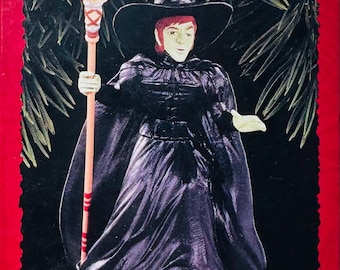 Peut inclure: Ornement Keepsake représentant la Méchante Sorcière de l'Ouest du Magicien d'Oz. La sorcière porte une robe et un chapeau noirs, tenant un balai. L'ornement est sur un fond rouge avec du texte.