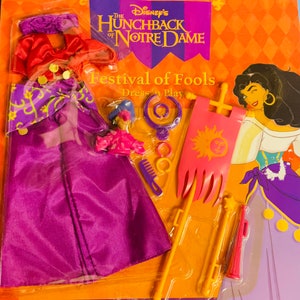 May include: A Disney's The Hunchback of Notre Dame Festival of Fools Dress-Up Play set. The set includes a purple dress, a red top, a yellow crown, a yellow comb, a pink banner, a yellow staff, a pink trumpet, and a doll with a purple dress and a blue hat.