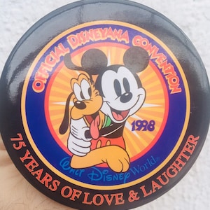 May include: A black and blue round button with a yellow sunburst design. Mickey Mouse and Pluto are hugging in the center. The text reads "Official Disneyana Convention 1998 75 Years of Love & Laughter Walt Disney World".