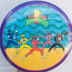 Könnte beinhalten: Ein runder, blauer und gelber Power Rangers-Aufkleber mit den Worten "Mighty Morphin Power Rangers" in einem weißen Kreis. Der Aufkleber zeigt fünf Power Rangers: den Roten Ranger, den Blauen Ranger, den Gelben Ranger, den Pinken Ranger und den Schwarzen Ranger. Jeder Ranger hält seine Waffe.