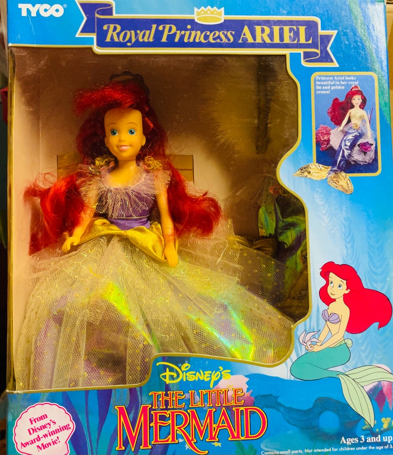 May include: A boxed Royal Princess Ariel doll from Disney's The Little Mermaid. The doll has red hair and a purple and yellow gown. The packaging includes the film's logo and images of Ariel. The box is labelled 'Ages 3 and up'.