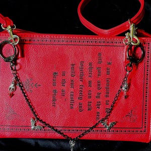 May include: A red book-shaped crossbody bag with a black chain and silver charms. The bag has a quote from Bram Stoker's "Dracula" printed on the front: "I am longing to be with you, and by the sea. Where we can talk together freely and build our castles in the air."