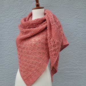 May include: A pink knitted shawl with a lacy pattern. The shawl is draped over a white mannequin.