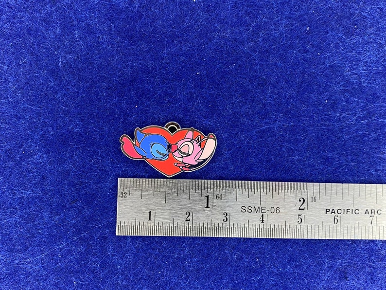 Puede incluir: Un pin en forma de coraz&oacute;n rojo con un Stitch azul y un Angel rosa de la pel&iacute;cula de Disney Lilo & Stitch bes&aacute;ndose. El pin mide aproximadamente 2,5 cm de ancho.
