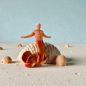 May include: A small merman figurine with a copper tail and tan skin, arms outstretched. The merman is positioned in front of seashells, with a light blue backdrop, creating a beach-like setting.