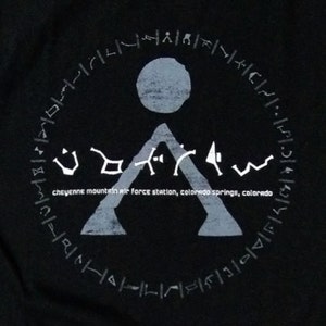 May include: A black t-shirt with a white graphic of a symbol with a circle and a triangle. The text "Cheyenne Mountain Air Force Station, Colorado Springs, Colorado" is printed below the symbol.