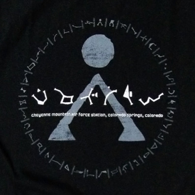 May include: A black t-shirt with a white graphic of a symbol with a circle and a triangle. The text "Cheyenne Mountain Air Force Station, Colorado Springs, Colorado" is printed below the symbol.