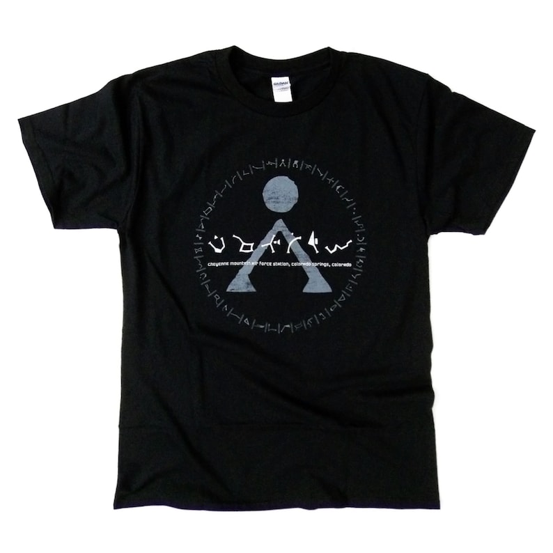 May include: Black t-shirt with a white graphic of a symbol with a circle around it. The symbol is a triangle with a circle on top and the text "UDOM4" underneath. The text "Cheyenne Mountain Air Force Station, Colorado Springs, Colorado" is written below the symbol.
