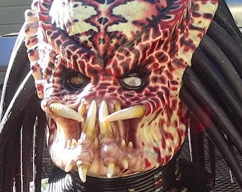 May include: A detailed Predator mask with a textured, reptilian appearance. The mask features a color palette of red, cream, and black, with prominent tusks and dreadlocks. The design is intricate, with a focus on the creature's menacing features.