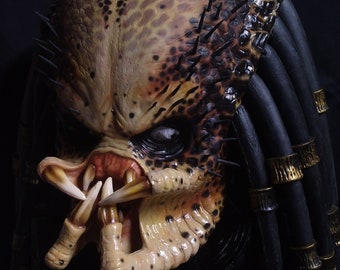 Op de afbeelding: Een gedetailleerd Predator-masker met een getextureerde, reptielachtige huid in tinten beige, bruin en zwart. Het masker heeft scherpe tanden, dreadlocks en een dreigende uitdrukking.