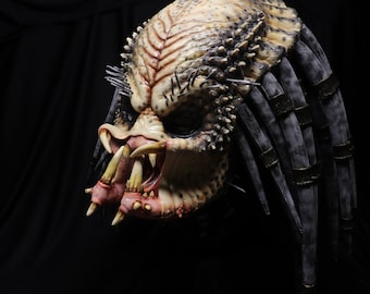 Puede incluir: Máscara detallada de Predator, con un bio-casco texturizado beige y marrón, púas afiladas y rastas. La máscara tiene mandíbulas grandes y amenazantes y dientes afilados, sobre un fondo oscuro.