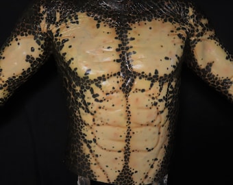 Op de afbeelding: Een prothetische torso met een reptielenhuidontwerp. De huid is een mix van bruin en zwart, met een patroon van donkere vlekken en schubben die de armen en borst bedekken. De borst heeft gedefinieerde buikspieren.