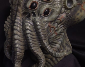 Puede incluir: Una máscara detallada, verde y gris con múltiples ojos y tentáculos. La máscara tiene una superficie texturizada y está diseñada para parecerse a una criatura. La máscara es probablemente para disfraces o cosplay.
