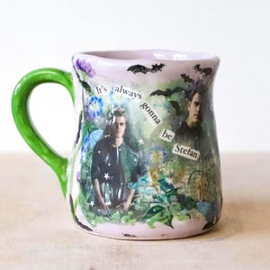 Puede incluir: Una taza de cerámica con asa verde y borde lila claro. La taza presenta imágenes de un hombre y el texto "It's always gonna be Stefan" con siluetas de murciélagos. También hay diseños florales y de estrellas.