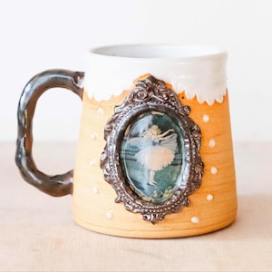 Könnte beinhalten: Handgefertigte Keramiktasse mit beigefarbenem Korpus, weißem Rand und dunkelbraunem Henkel. Die Tasse zeigt ein gerahmtes Bild einer Ballerina und kleine weiße Punkte. Ein einzigartiges Trinkgefäß.