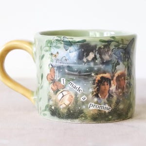 Peut inclure: Une tasse en céramique vert clair avec une anse jaune, ornée d'un collage imprimé représentant deux personnages, une bague dorée, des papillons et le texte I made a promise. La tasse présente un bord organique irrégulier et des motifs de vigne.