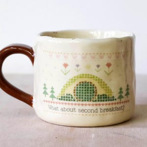 May include: Cream-colored ceramic mug with a brown handle. The mug features a whimsical design with a green and yellow arched structure, trees, and the text "What about second breakfast?" in a playful font.