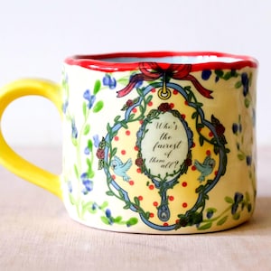 Könnte beinhalten: Keramiktasse mit gelbem Henkel und Rand, verziert mit Blumen- und Vogelmotiven. Die Tasse trägt den Text "Who's the fairest of them all?" in einem dekorativen ovalen Spiegeldesign. Der Innenrand ist rot.