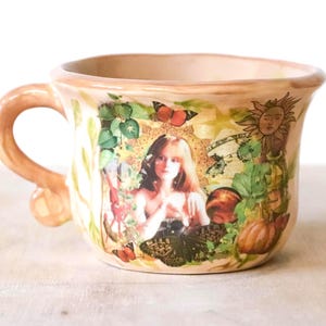 Puede incluir: Una taza de cerámica beige con un asa grande, decorada con un retrato de mujer, mariposas, un sol y follaje. La taza tiene un borde ancho y una base redondeada. El diseño es en tonos verdes, naranjas y marrones.
